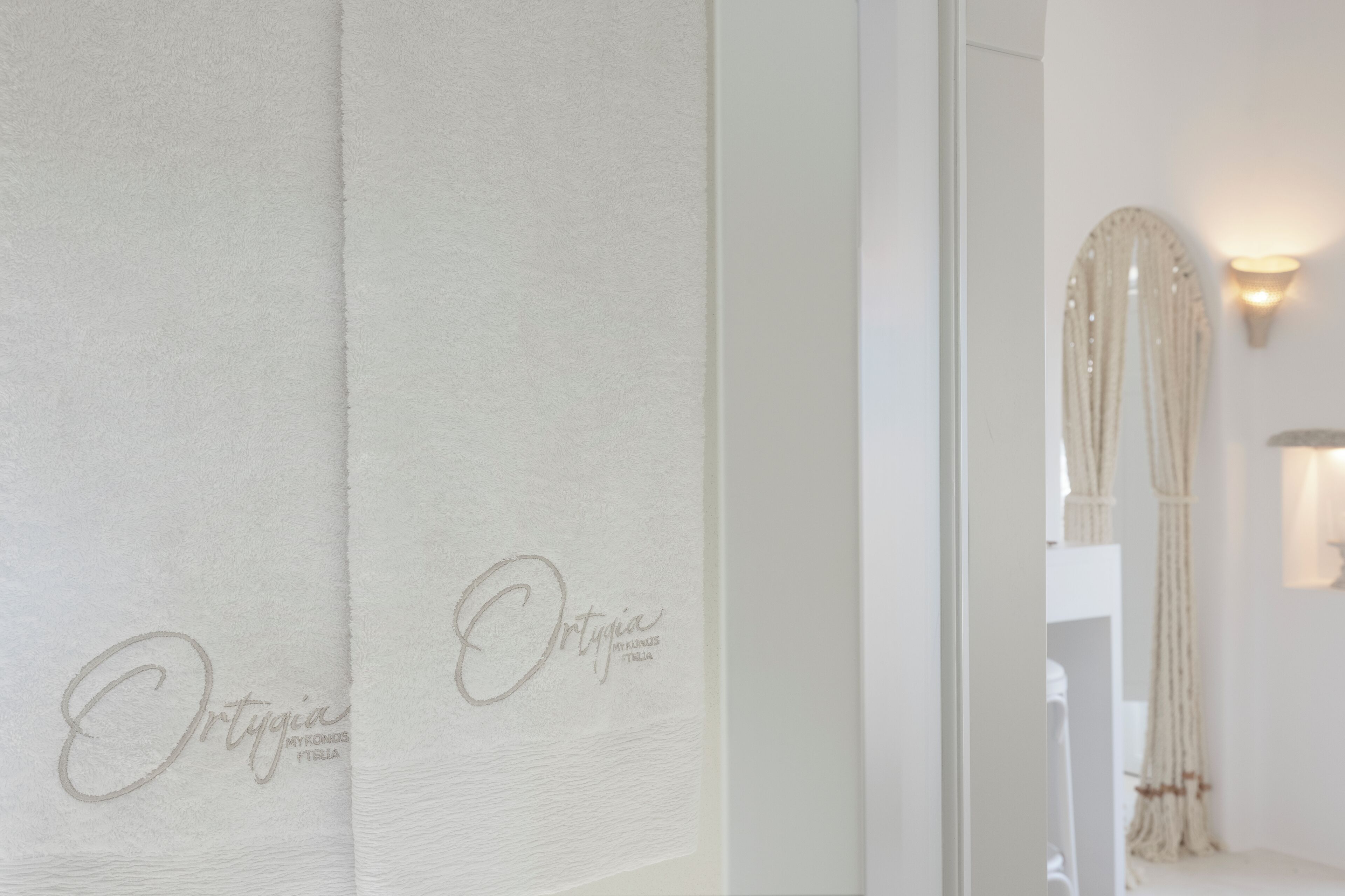 A hotel room with a white curtain featuring the name "Ortigia" and a wall-mounted light fixture.