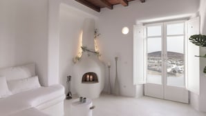 Una sala de estar moderna con un sofá blanco, una chimenea con dos velas, jarrones altos y un ventanal con vistas a un pueblo.