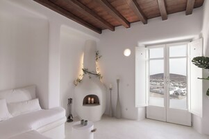Uma sala de estar moderna com um sofá branco, uma lareira com duas velas, vasos altos e uma janela grande com vista para uma cidade.