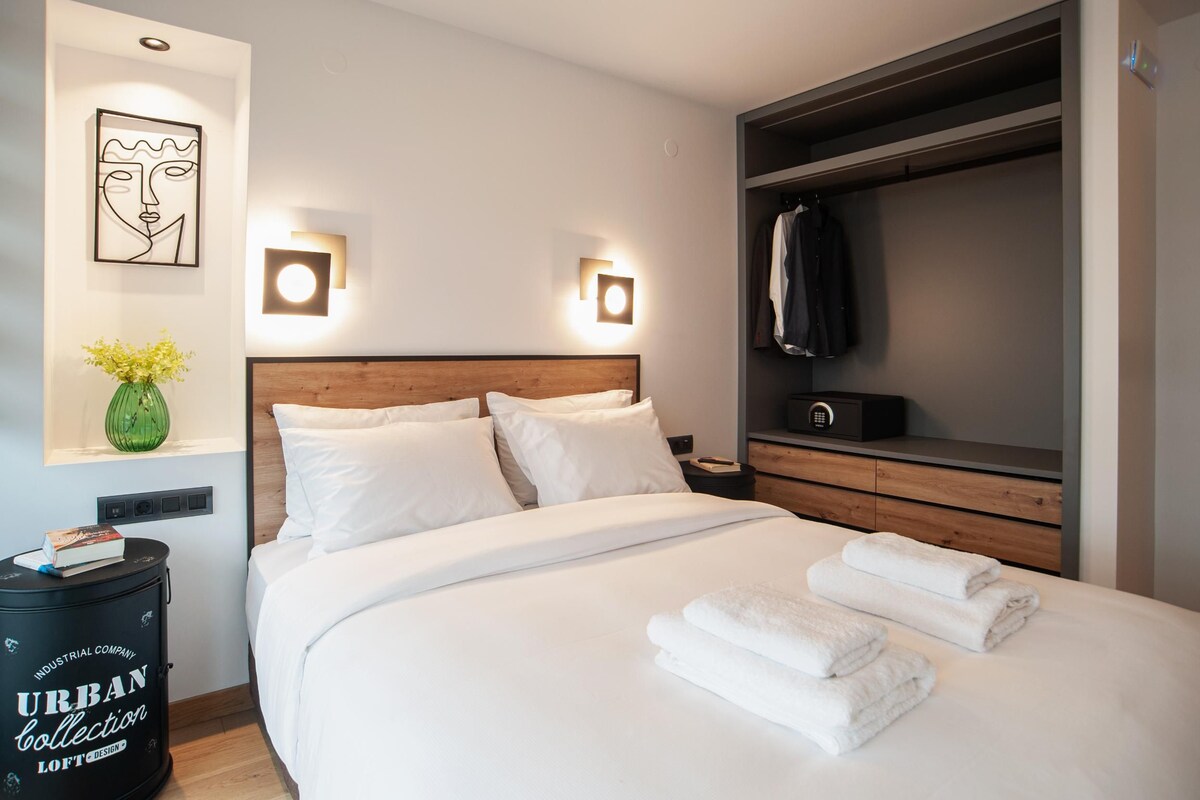 A modern hotel room with a large bed, a wooden headboard, a wardrobe, and a bedside table with a lamp.