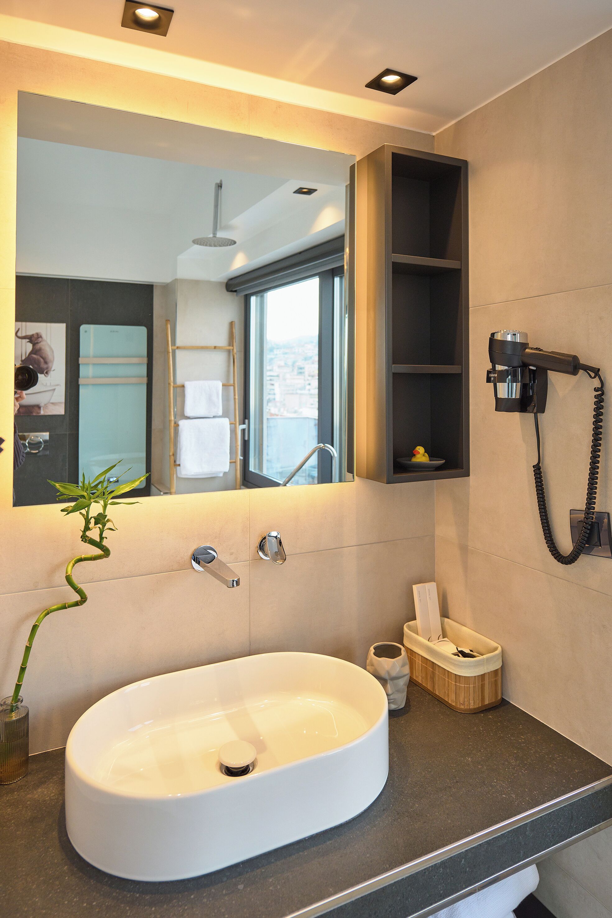 A modern bathroom with a large oval sink, a mirror, a hairdryer, and a small shelf with a towel and a decorative item.