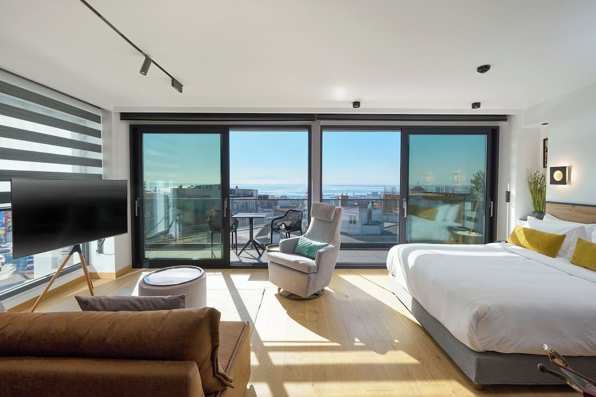 A modern hotel room with a large bed, a flat-screen TV, a seating area with a chair and table, and a balcony with a view of the city.