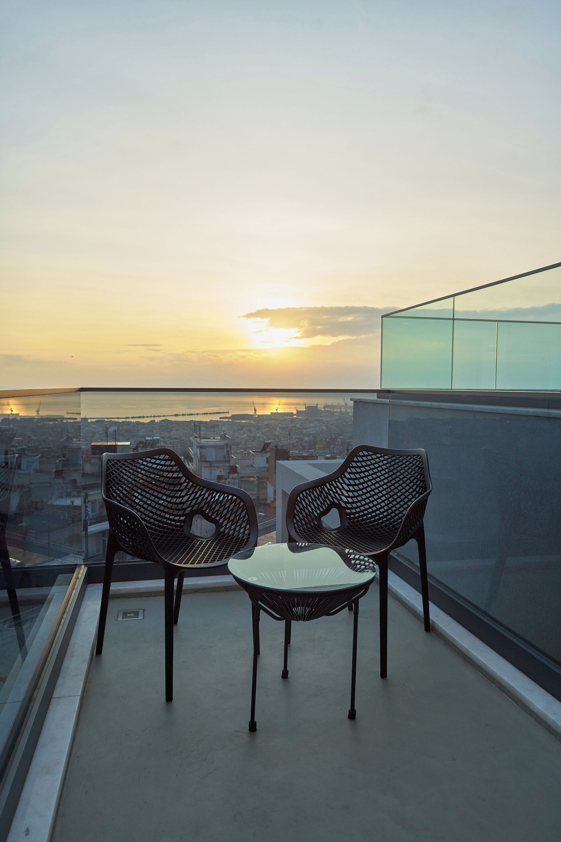 A balcony with two chairs and a table, overlooking a cityscape at sunset.