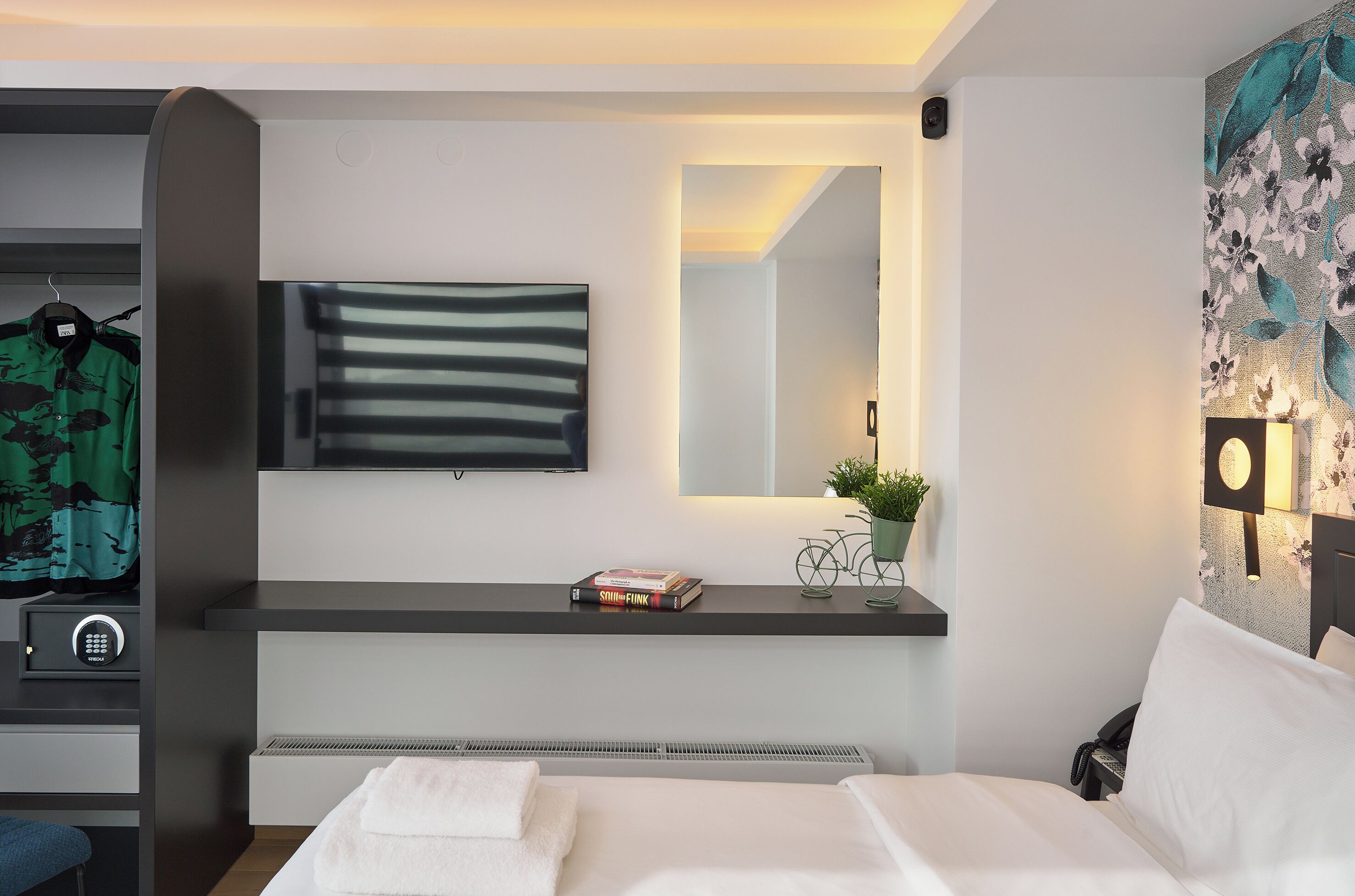 Et moderne hotelværelse med seng, sengebord med lampe, ophængt fjernsyn og spejl på væggen.