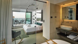 A modern hotel room with a large bed, a glass partition, a balcony with outdoor seating, and a bathroom with a large mirror and sink. - SIGMA Suites Hotel (Neapoli-Sykies)