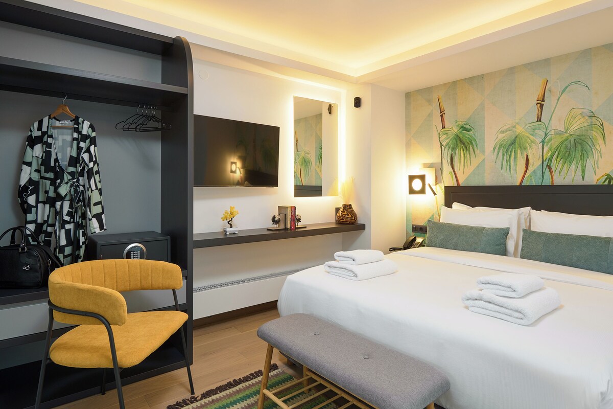 A hotel room with a bed, a bench, a chair, a wardrobe, a mirror, and a wall with a tropical mural.