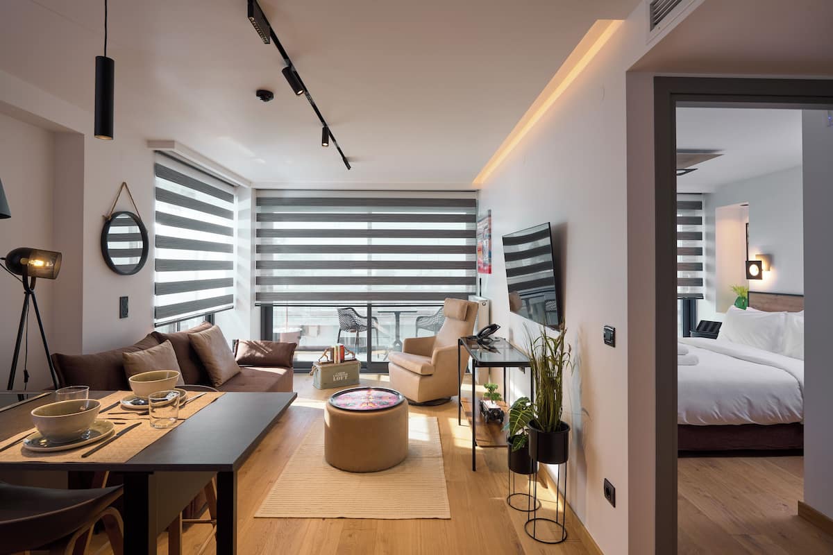 A modern living room with a sofa, armchair, dining table, and a large window with blinds.