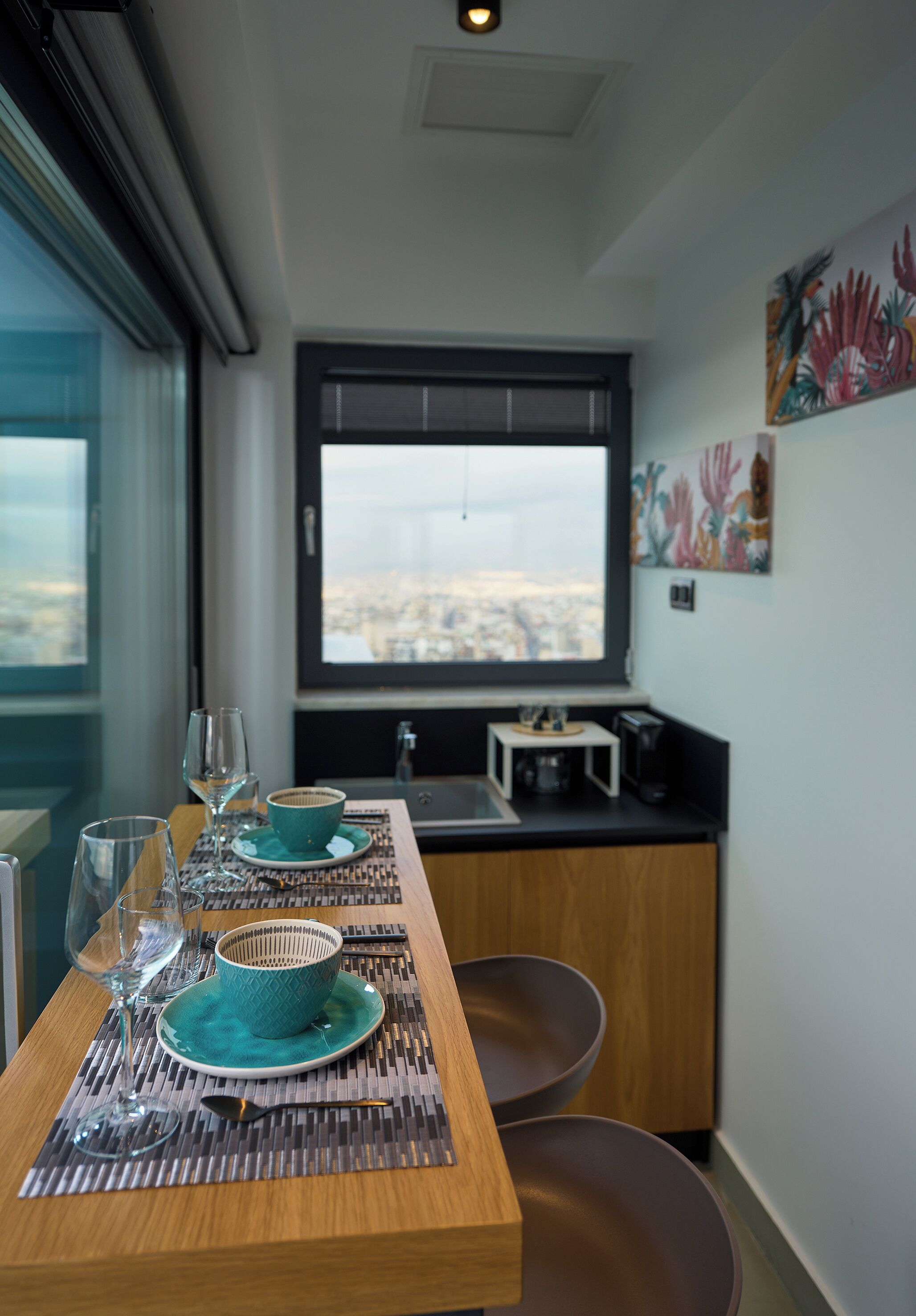 Un comedor con una mesa lista para dos personas, una ventana con vistas a un paisaje urbano y una cocina con electrodomésticos modernos.