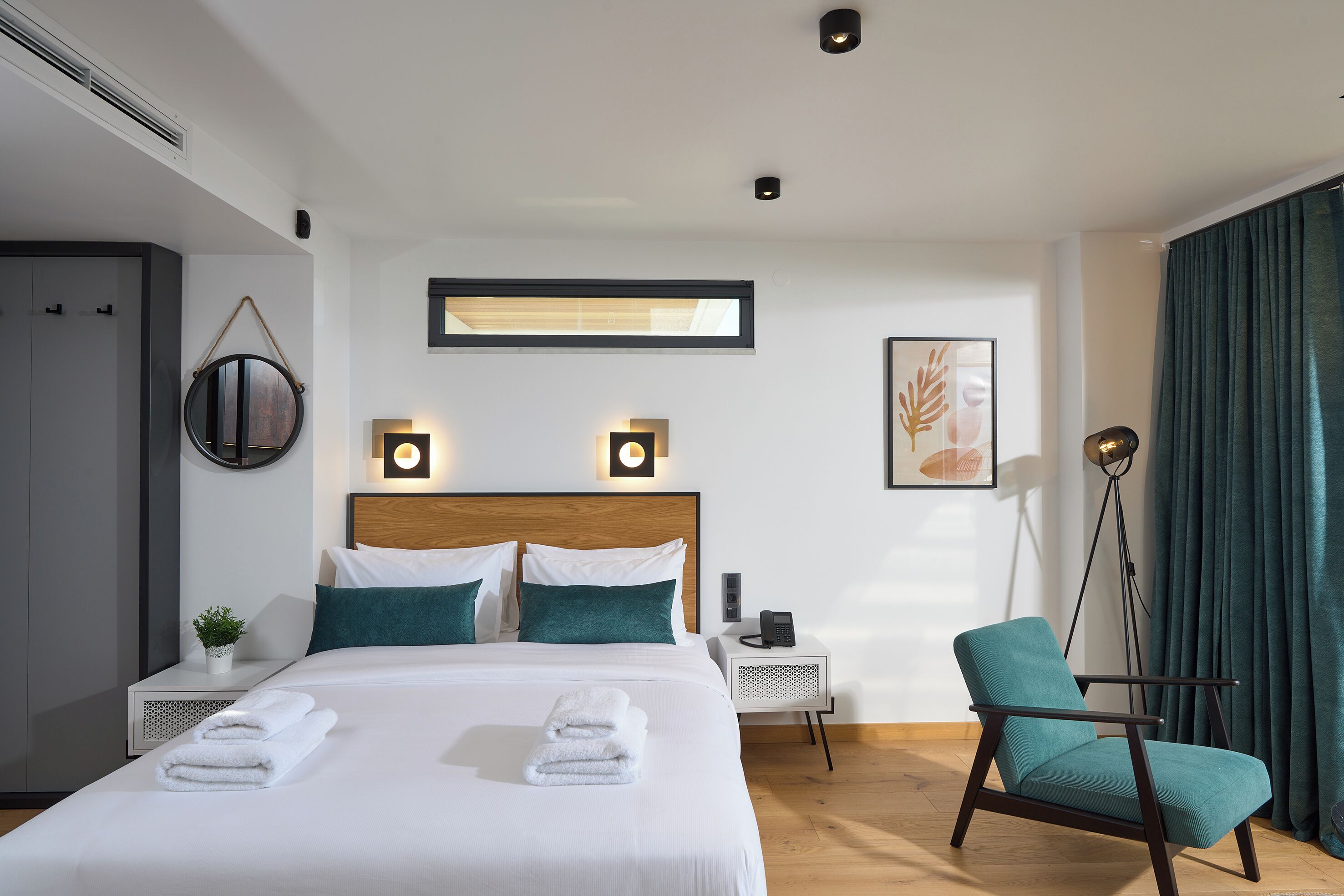 Et moderne hotelværelse med en pænt redt seng, et natbord med lampe, en stol og et stort vindue med gardiner.