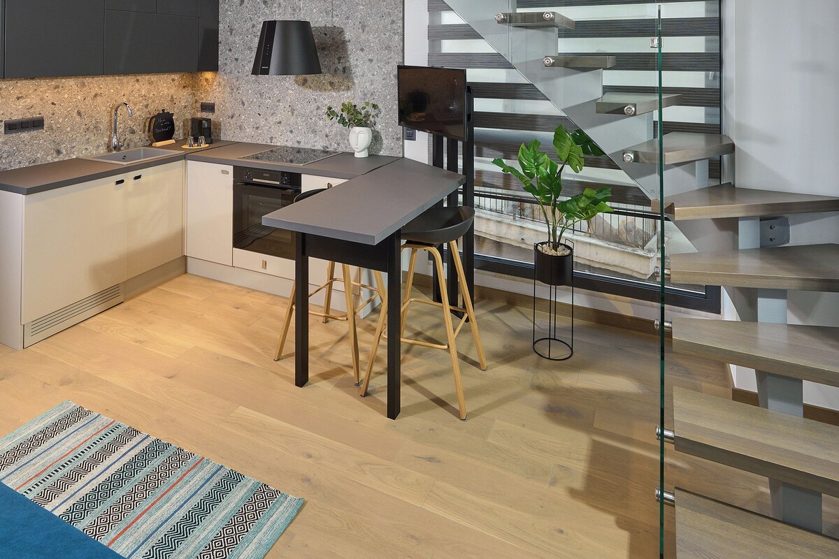 A modern kitchen with a built-in oven, a dining area with a table and chairs, and a staircase with glass railings leading to an upper level.