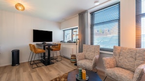 Ein modernes Wohnzimmer mit Flachbildfernseher, zwei Sesseln, einem Couchtisch und einem Essbereich mit kleinem Tisch und Stühlen.