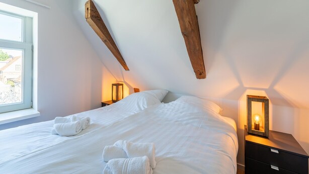 Ein Schlafzimmer mit einem großen Bett, Holzbalken, zwei Nachttischen mit Lampen und einem Fenster.