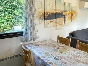 A dining area with a table covered in a patterned tablecloth, a window with a view of greenery, and a sofa in the background.