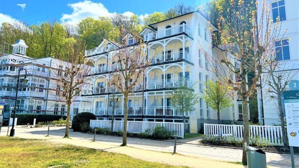 Un hotel de varios pisos con balcones, una cerca blanca y árboles en primer plano.