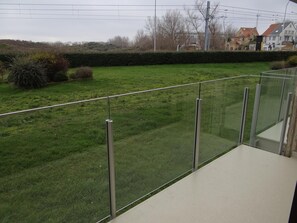 Ein Balkon mit Metallgeländer, der auf eine Rasenfläche mit Büschen und einer Hecke blickt.