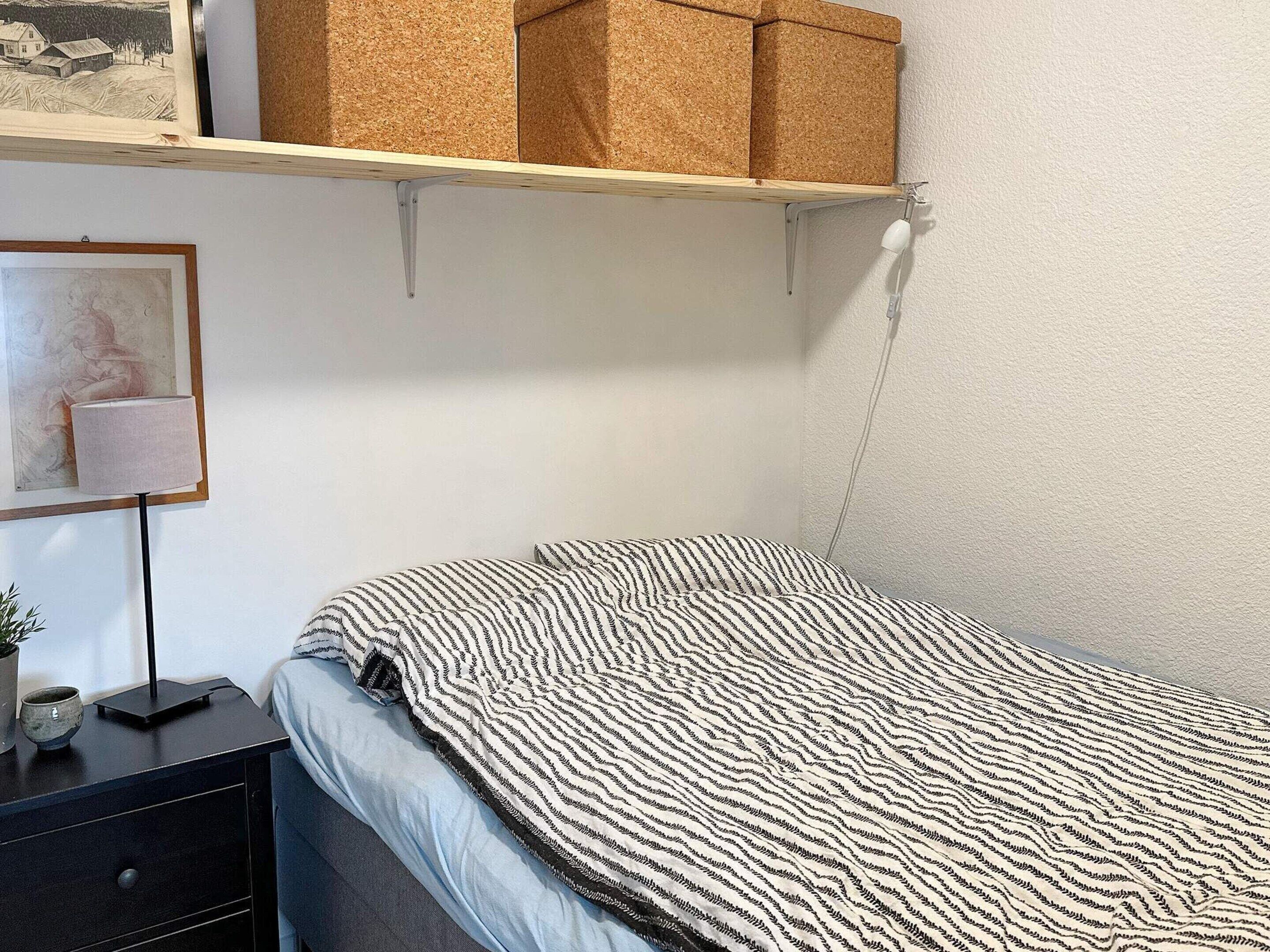 A bedroom with a bed, a nightstand, a lamp, a framed picture, and storage boxes on a shelf.