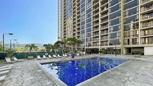 A hotel pool area with a clear view of the pool, lounge chairs, and a tall building in the background. - Ocean & Diamond Head Views! Full Kitchen, Lanai, FREE Parking, Wi-Fi & TV! (Honolulu)