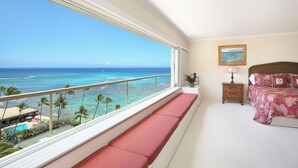 A bedroom with a balcony overlooking a beach, a bed with floral bedding, and a nightstand with a lamp. - Breathtaking Ocean Views from Quiet Beachfront Condo! Free Parking & Wi-Fi! (Honolulu)