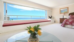 A bedroom with a large window overlooking a beach, a bedside table with a lamp, a bed with floral bedding, and a vase with flowers on a glass table. - Breathtaking Ocean Views from Quiet Beachfront Condo! Free Parking & Wi-Fi! (Honolulu)