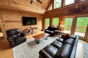L'intérieur d'un chalet en bois, avec un téléviseur, des meubles en cuir et de grandes fenêtres.