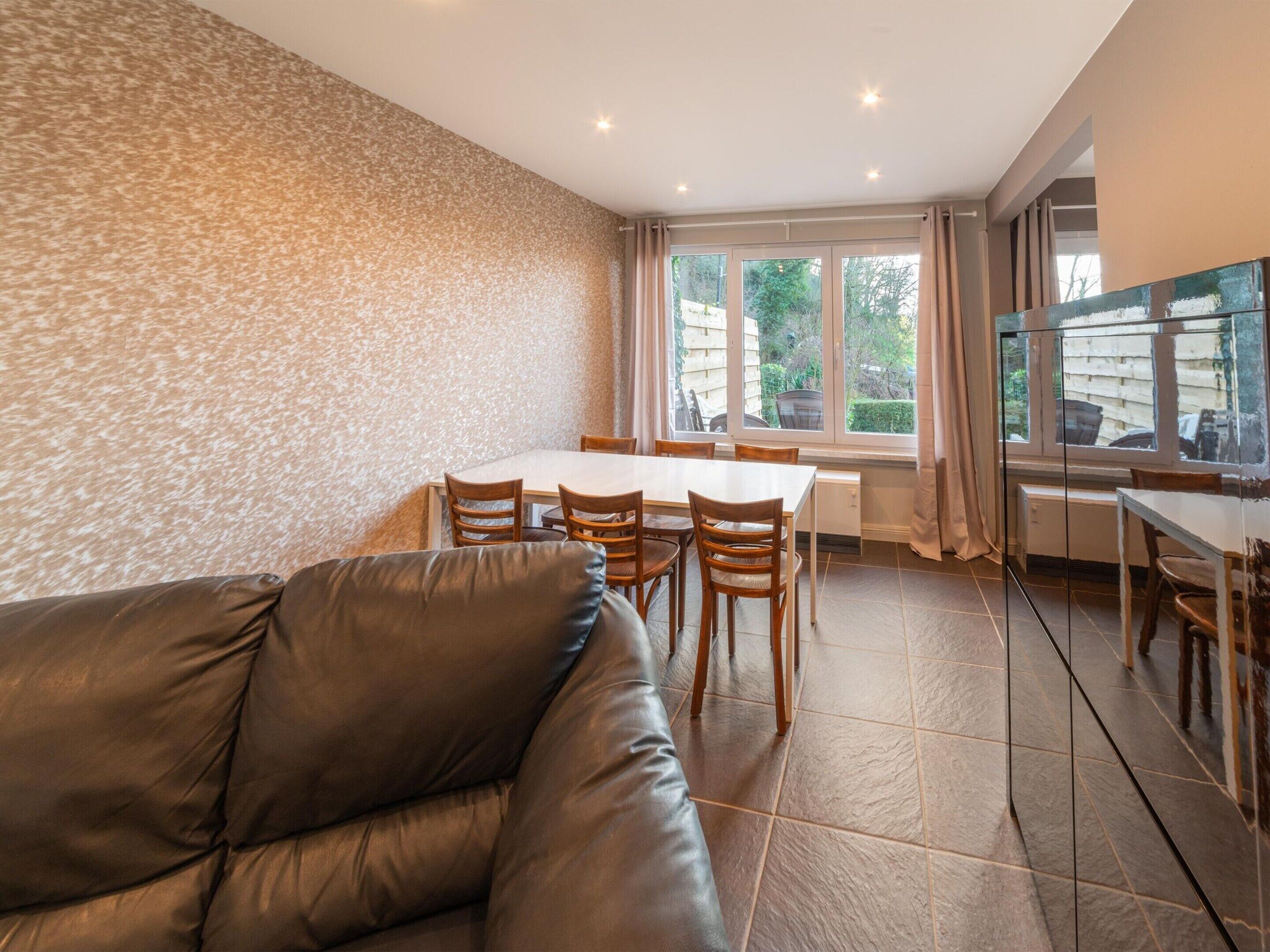 Uma sala de estar moderna com um sofá de couro, uma mesa de jantar com cadeiras e grandes janelas com vista para o exterior.