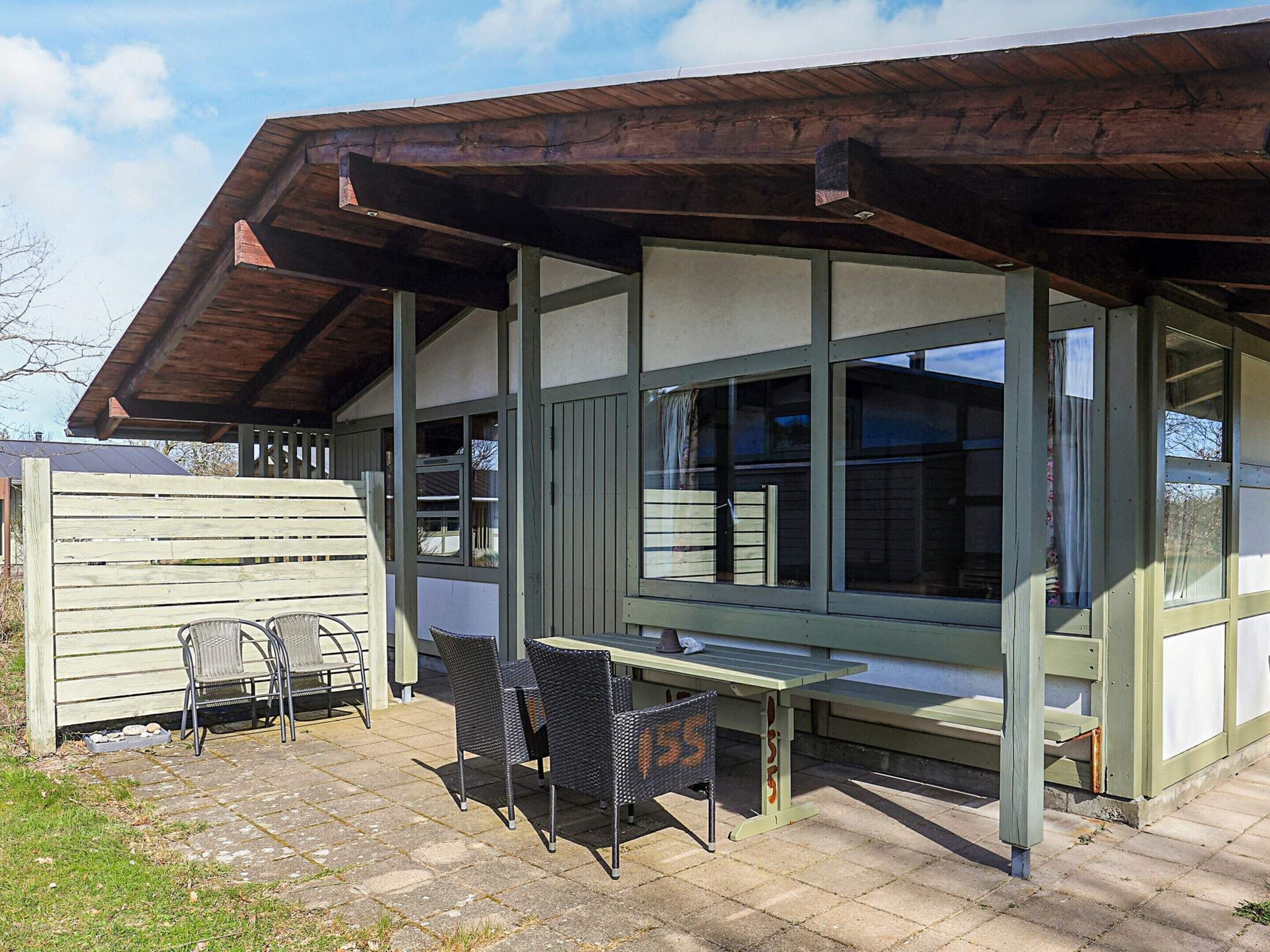 Een overdekt terras met een tafel en stoelen, een houten hek en een gebouw met grote ramen.