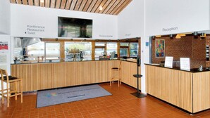 Zona de recepción con mostrador de madera, escritorio de recepción y señalización para conferencias, restaurante y baños.