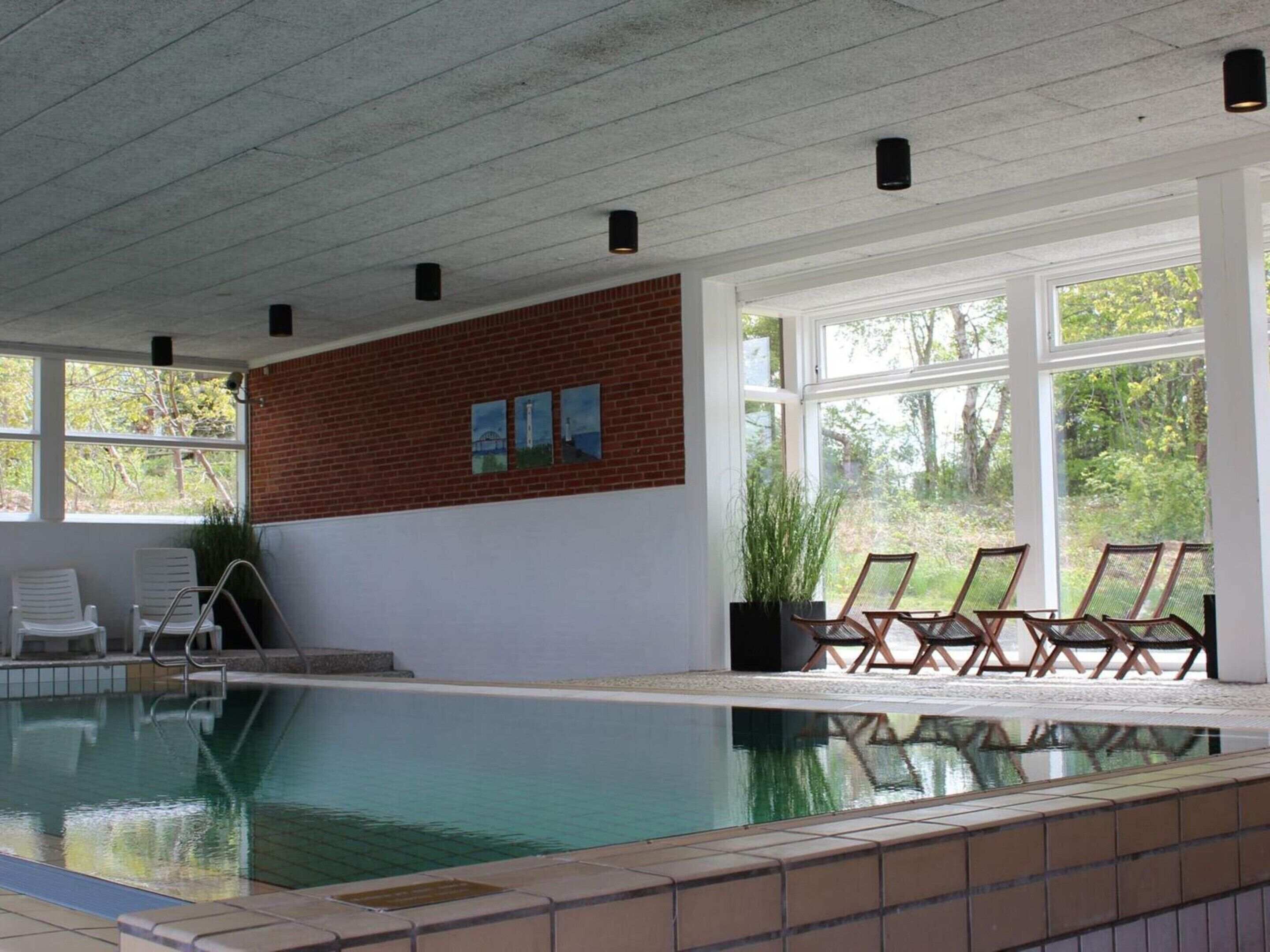 An indoor swimming pool with clear glass walls, a brick accent wall, and several lounge chairs.