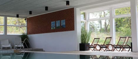 An indoor swimming pool with clear glass walls, a brick accent wall, and several lounge chairs.