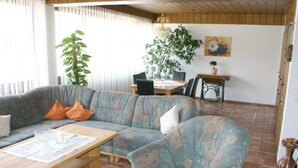 Uma sala de estar com sofá, mesa de centro de madeira, mesa de jantar com cadeiras e um vaso com planta.