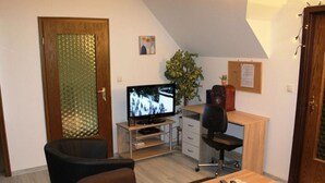 Uma sala de estar pequena com televisão, escrivaninha, cadeira e uma planta.