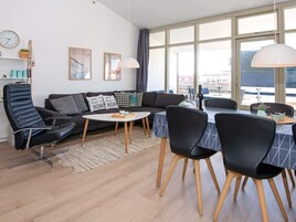 Una sala de estar moderna con un sofá, sillas, una mesa de centro y una mesa de comedor. Hay ventanas amplias, un reloj y cuadros en la pared.