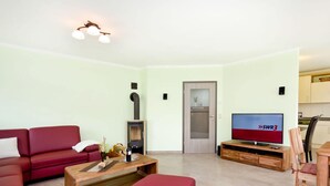 A living room with a red sofa, a wooden coffee table, a red ottoman, a fireplace, a television, and a kitchen area in the background. - Apartment in Tisens (Putbus)
