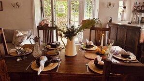 A dining room with a wooden table set for a meal, surrounded by chairs. There are windows with a view of greenery outside. - Charming Holiday Cottage in Devon - Country Views (Tiverton)