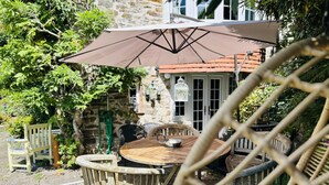 An outdoor seating area with a round wooden table and chairs, a large umbrella, and a stone building in the background. - Charming Holiday Cottage in Devon - Country Views (Tiverton)