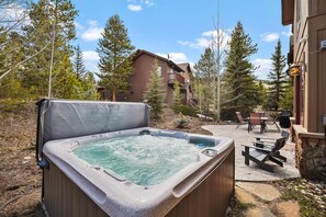 Un jacuzzi en un patio trasero con un suelo de piedra, rodeado de árboles y un edificio de madera.