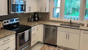 A modern kitchen with white cabinets, stainless steel appliances, a granite countertop, and a window with a view of greenery. - Serenity Home - Forest, Fun, and Adventure (Brevard)