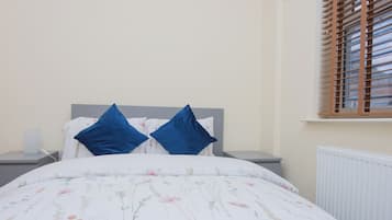 A neatly made bed with a floral-patterned duvet, two blue pillows, and a grey headboard. There is a bedside table with a lamp and a power outlet. A window with wooden blinds is visible in the background.