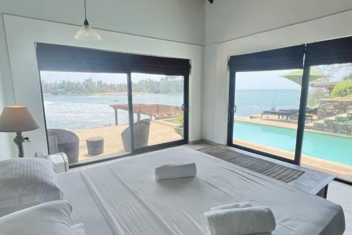 Een slaapkamer met een groot bed, uitzicht op een zwembad en terras, en een raam met uitzicht op de zee.