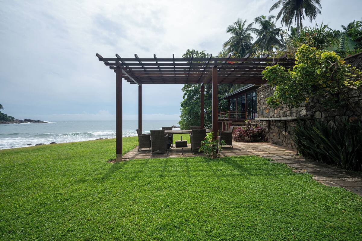 Een terras met een pergola dat uitkijkt over de oceaan, een stenen muur en weelderige begroeiing.