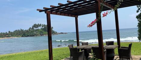 A wooden pergola with wicker furniture overlooking a beach with waves and palm trees.