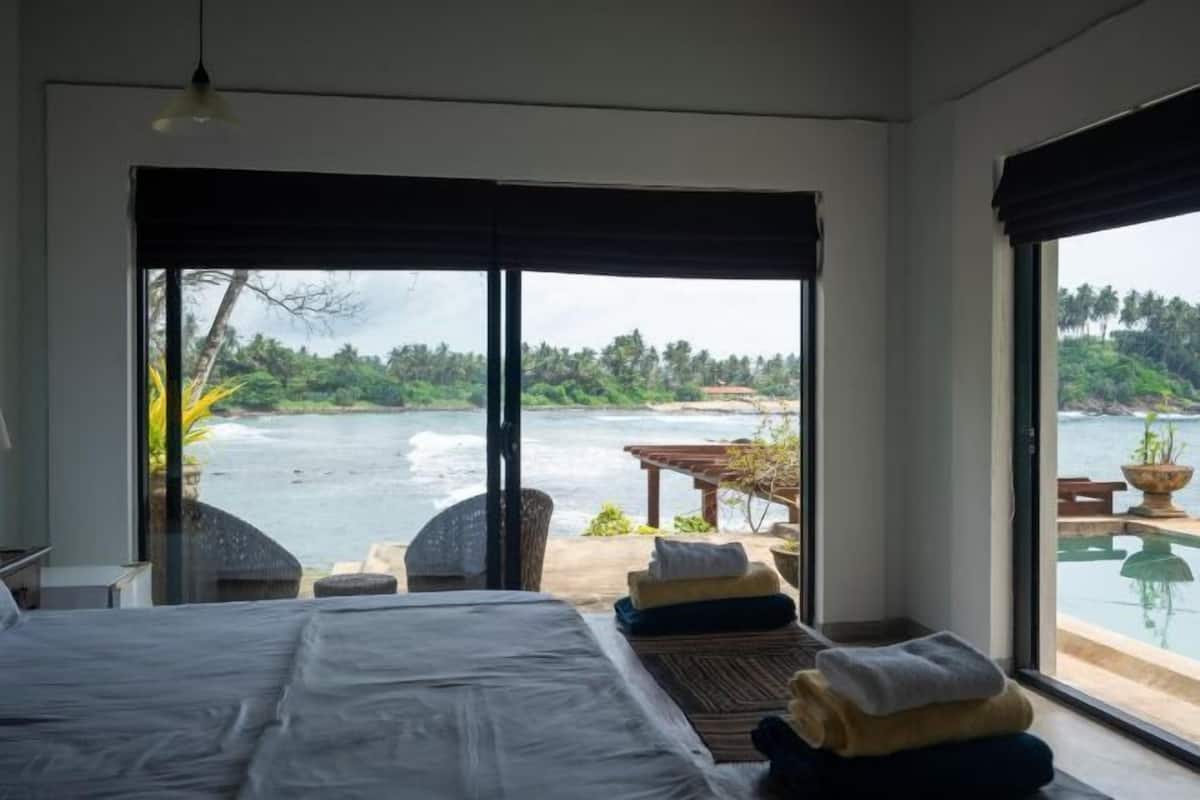 Een slaapkamer met een groot bed, uitzicht op een waterpartij en een terras met zitplaatsen.