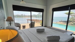 Een slaapkamer met een groot bed, een nachtkastje, een lamp en uitzicht op een zwembad en strand.