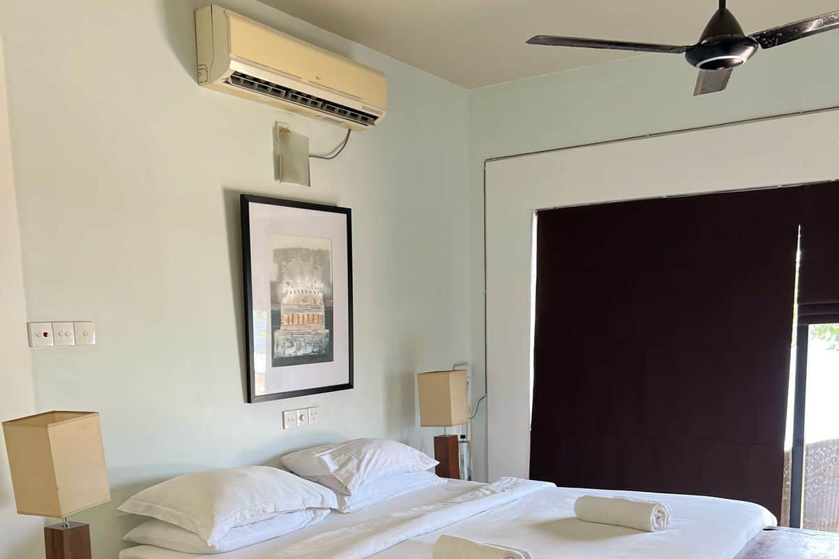 Een hotelkamer met een bed, een plafondventilator, een airconditioning, een ingelijste foto, een nachtkastje, een lamp en een raam met gordijnen.