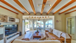 A spacious living room with a large TV, wooden beams, a sofa, and a view through large windows. - Casa Palmeras - Moraira (Moraira)