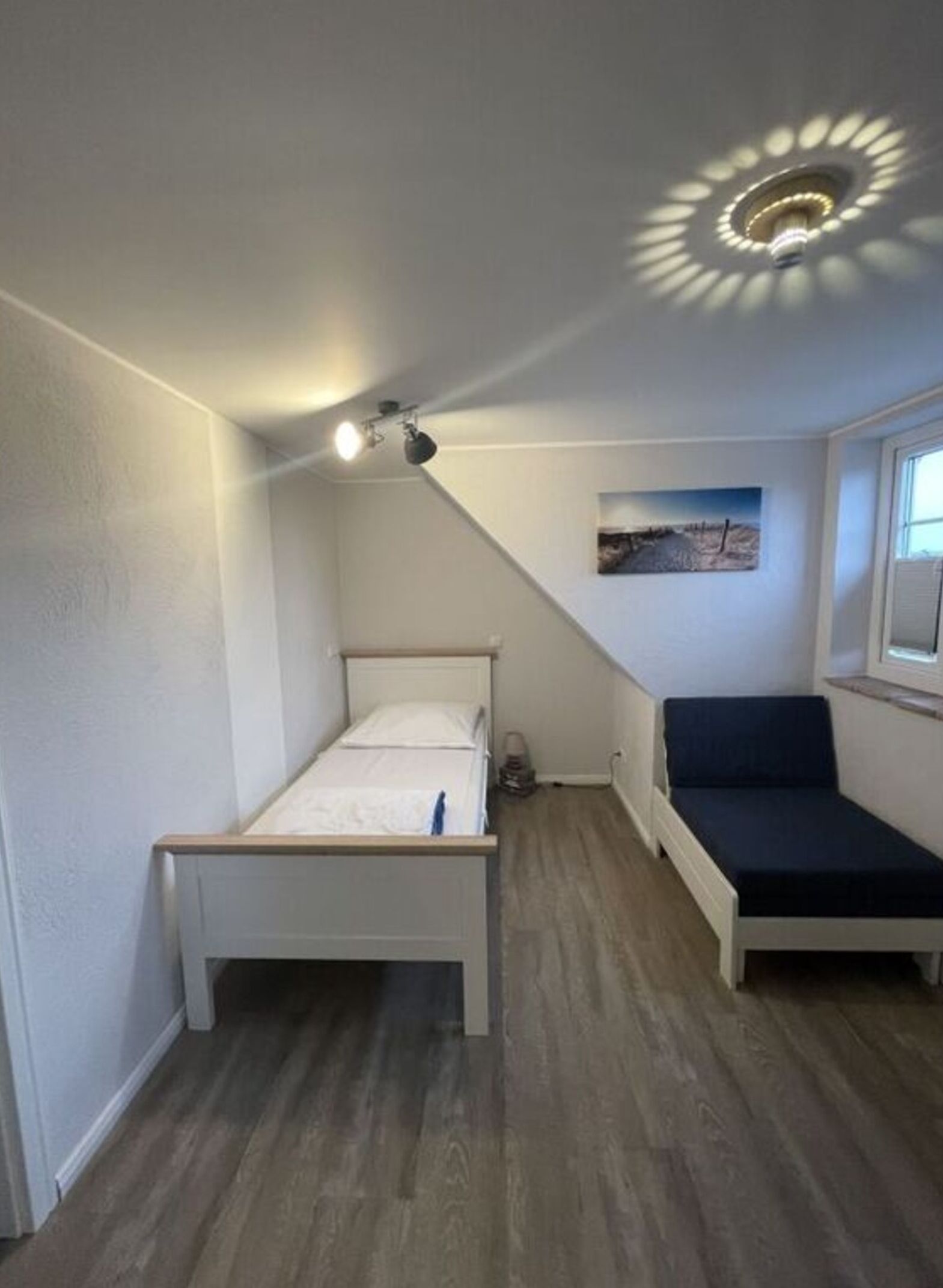 A small, single-room apartment with a bed, a bench, and a ceiling light fixture.