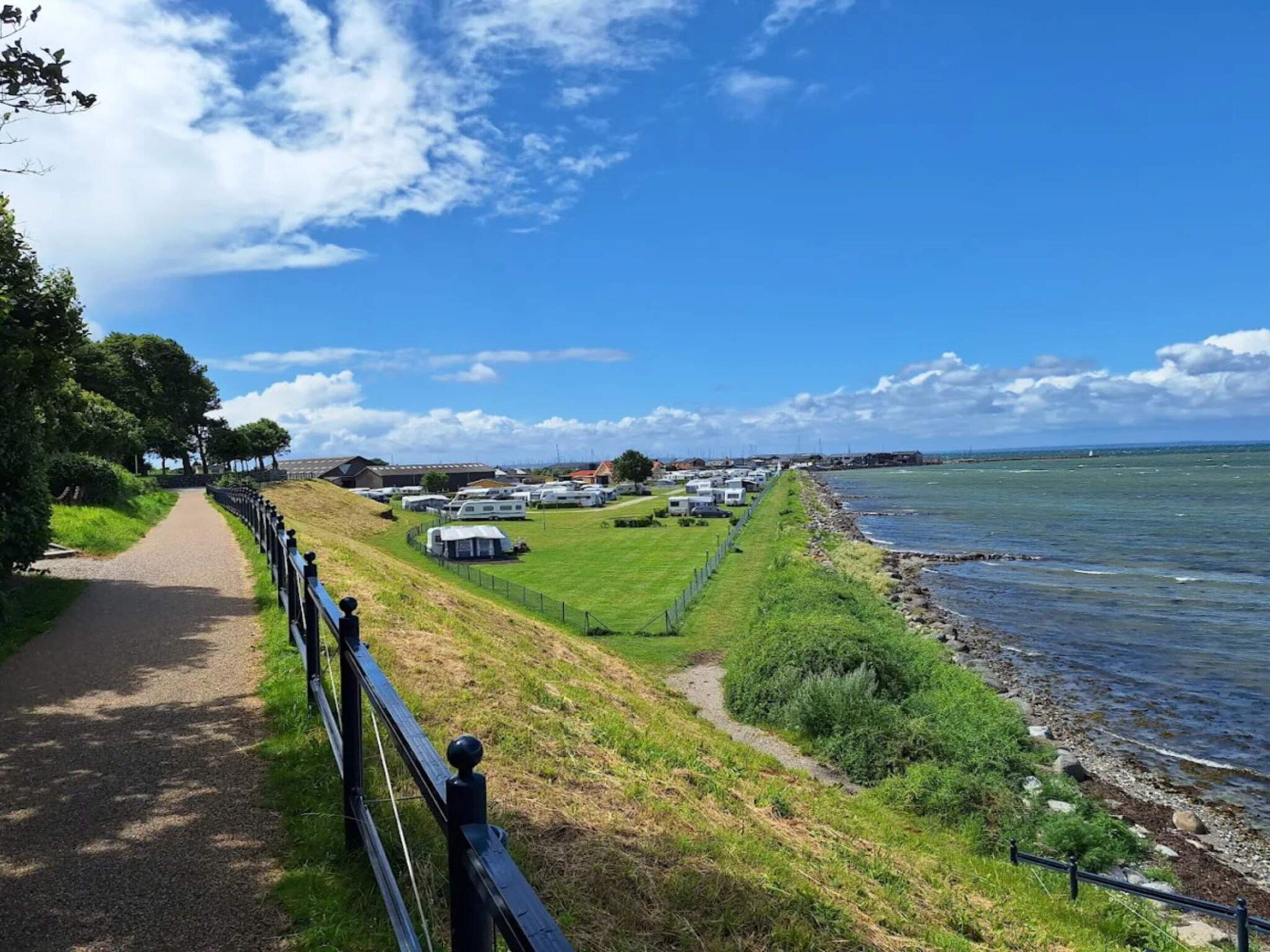 Un sentier côtier bordé d’une clôture, avec des zones herbeuses et une rangée de maisons mobiles au bord de la mer.