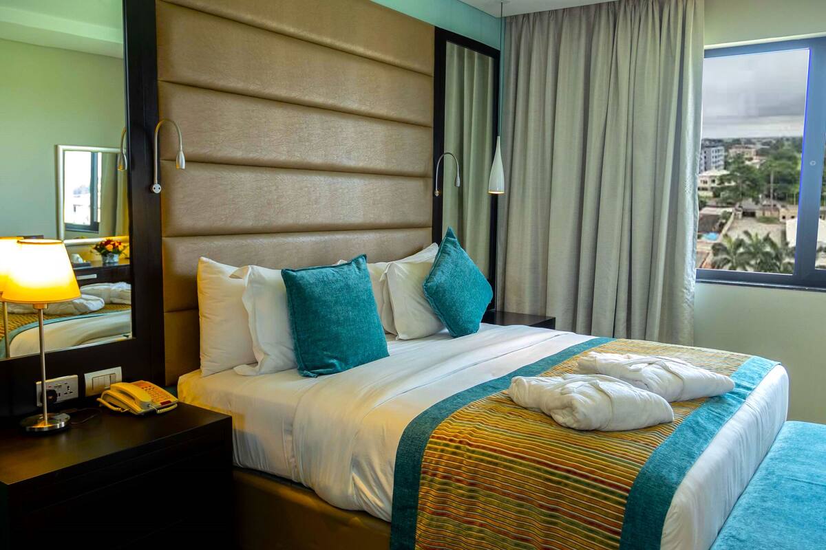 Una camera d'albergo con un letto grande, un comodino con lampada, un telefono e una finestra con tende.