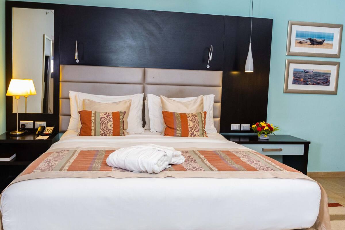 Camera d'albergo con un letto grande, un comodino con una lampada, un tavolino da notte con un vaso di fiori e una parete decorata con quadri.