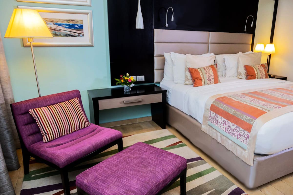 Una camera d'albergo con un letto, un comodino, una sedia, una panca, una lampada e una televisione a parete.